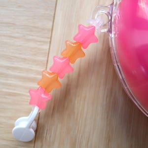 May include: A decorative charm featuring a vertical string of five alternating pink and orange star-shaped beads. The strand is attached to a clear plastic loop on a bright pink spherical object and terminates in a small white plastic clip.