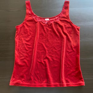 Puede incluir: Una camiseta sin mangas de terciopelo rojo con cuello redondo y delicado ribete de encaje. La parte superior presenta un pequeño detalle de lazo en el centro del escote. La tela tiene una textura suave y afelpada.