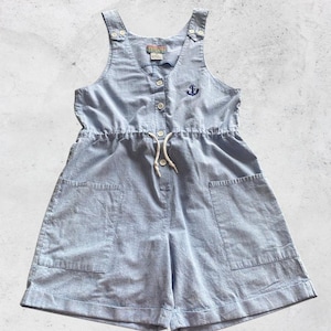 May include: Light blue romper with adjustable straps and a drawstring waist. The romper features a small embroidered anchor on the chest and two front pockets. The fabric has a subtle striped pattern. Buttons run down the front of the romper.