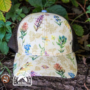 May include: A trucker hat featuring a cream-colored front panel with a colorful botanical print of various wildflowers and butterflies, paired with dark brown mesh side and back panels and a light blue button on the top crown.