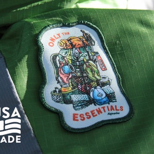 May include: A green patch with a white border and a cartoon illustration of a backpack filled with camping gear. The text "Only the Essentials" is printed in white on a red background. The text "Alpacka" is printed in white on a black background.