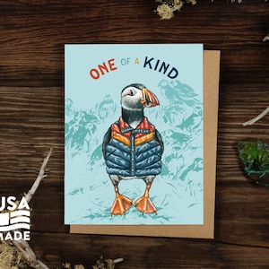 May include: A greeting card with a blue background featuring a cartoon puffin wearing a blue and orange puffer jacket. The text "ONE OF A KIND" is printed in orange and yellow letters on the card.