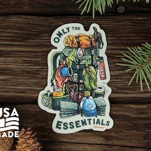May include: A sticker with a cartoon illustration of a green backpack filled with camping essentials. The text "ONLY THE ESSENTIALS" is written in a white banner at the bottom of the sticker. The text "Alpinacho" is written in smaller font below the banner. The sticker is on a wooden background.