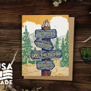 May include: A greeting card with a rustic design. The card features a signpost with directional signs that read "I'D BE LOST WITHOUT YOU." The background includes a mountain scene with trees and an orange sky. The words "USA MADE" are also visible.