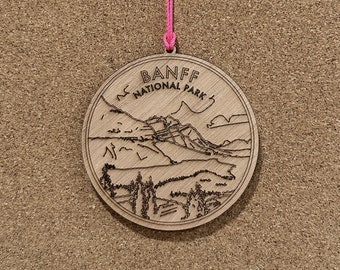 May include: A wooden ornament with a pink string. The ornament is round and features a black and white illustration of a mountain range with the text "BANFF NATIONAL PARK".