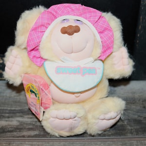May include: A plush yellow and pink teddy bear wearing a pink and white bonnet and a blue and white bib. The bib says "sweet pea". The bear is sitting on a wooden surface. The bear has a brown nose and purple eyes.