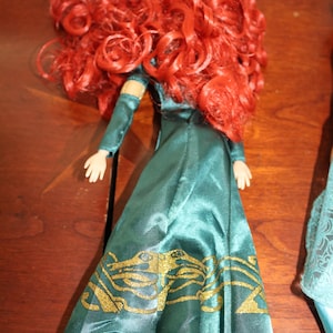 Puede incluir: Una mu&ntilde;eca con pelo largo y rizado rojo y un vestido verde azulado con detalles dorados. El vestido de la mu&ntilde;eca tiene mangas largas y un patr&oacute;n dorado detallado en la falda. La mu&ntilde;eca est&aacute; sobre una superficie de madera.