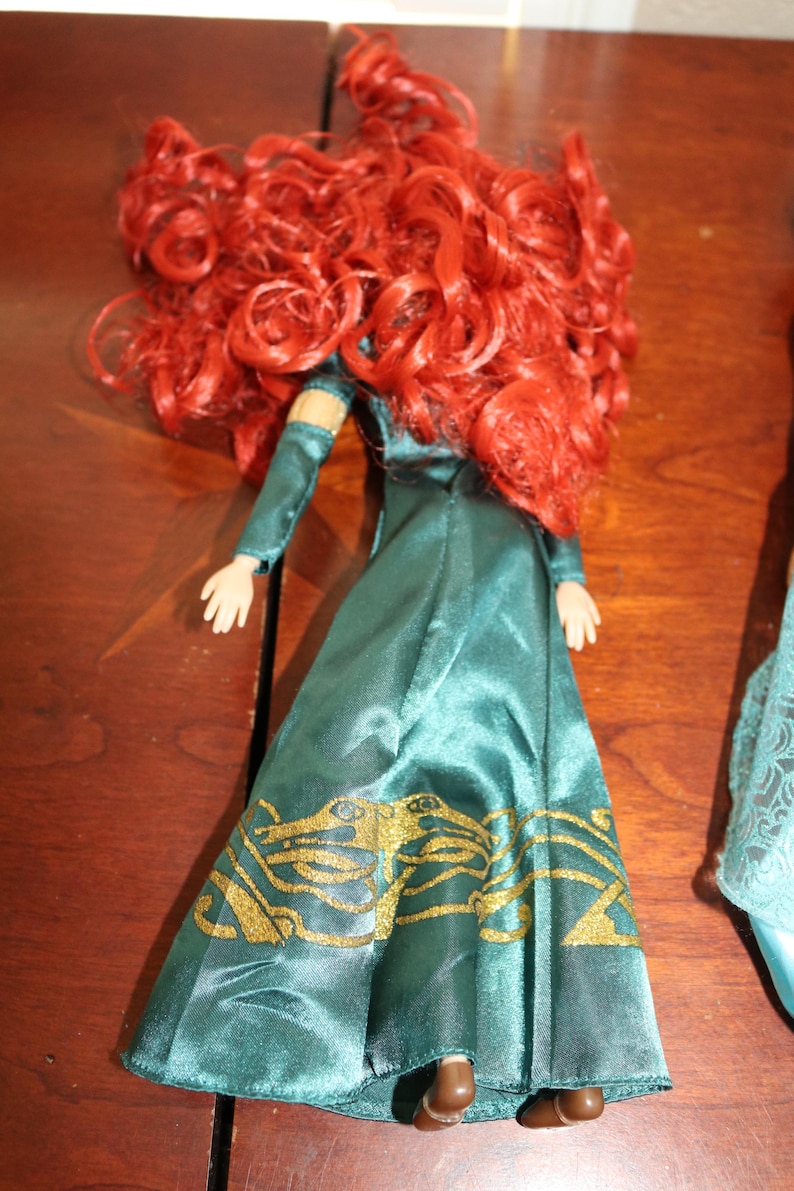 Puede incluir: Una mu&ntilde;eca con pelo largo y rizado rojo y un vestido verde azulado con detalles dorados. El vestido de la mu&ntilde;eca tiene mangas largas y un patr&oacute;n dorado detallado en la falda. La mu&ntilde;eca est&aacute; sobre una superficie de madera.