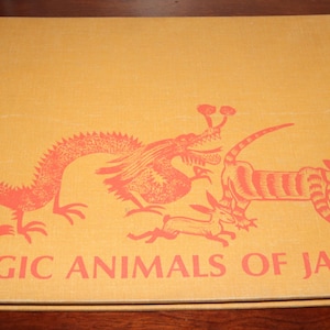 May include: A hardcover book titled "MAGIC ANIMALS OF JAPAN" with a tan cover. The cover features a red illustration of a dragon, a tiger-like creature, and other mythical animals. The book is on a dark wood surface.