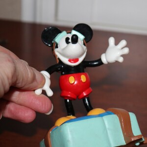 May include: A Mickey Mouse figurine with black ears, a black body, red shorts, and white gloves. The figurine is standing on a light blue suitcase with brown accents. The figure is wearing a light blue visor.
