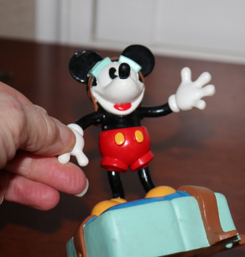 May include: A Mickey Mouse figurine with black ears, a black body, red shorts, and white gloves. The figurine is standing on a light blue suitcase with brown accents. The figure is wearing a light blue visor.