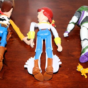 Puede incluir: Tres figuras de acci&oacute;n de personajes de la serie de pel&iacute;culas animadas. Woody, con un atuendo de vaquero, Jessie, con un atuendo de vaquera, y Buzz Lightyear, con un traje espacial, se muestran con una peque&ntilde;a figura de gato amarillo.