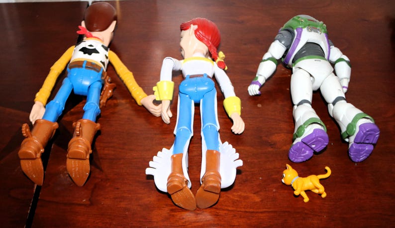 Puede incluir: Tres figuras de acci&oacute;n de personajes de la serie de pel&iacute;culas animadas. Woody, con un atuendo de vaquero, Jessie, con un atuendo de vaquera, y Buzz Lightyear, con un traje espacial, se muestran con una peque&ntilde;a figura de gato amarillo.