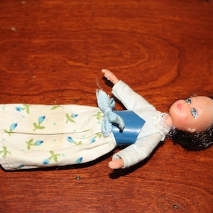 May include: A vintage doll with dark hair and blue eyes, wearing a white top, a blue bodice, and a floral-patterned skirt. The doll is lying on a wooden surface.