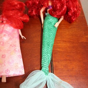 Puede incluir: Dos mu&ntilde;ecas de sirena. Una mu&ntilde;eca tiene una cola verde escamada y pelo rojo. La otra mu&ntilde;eca lleva un vestido rosa con un estampado floral y tiene el pelo rojo. Las mu&ntilde;ecas est&aacute;n sobre una superficie marr&oacute;n.