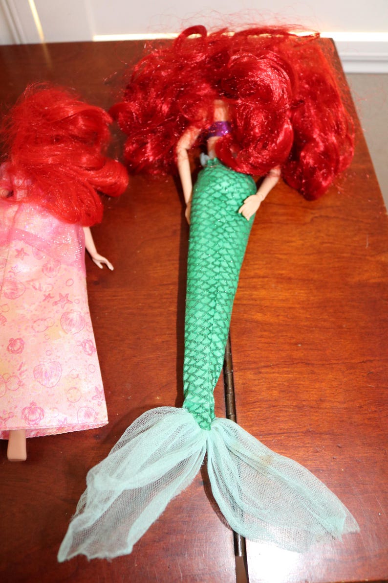 Puede incluir: Dos mu&ntilde;ecas de sirena. Una mu&ntilde;eca tiene una cola verde escamada y pelo rojo. La otra mu&ntilde;eca lleva un vestido rosa con un estampado floral y tiene el pelo rojo. Las mu&ntilde;ecas est&aacute;n sobre una superficie marr&oacute;n.