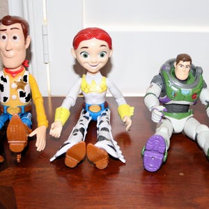 Puede incluir: Tres figuras de personajes de Toy Story: Woody, Jessie y Buzz Lightyear, junto con una peque&ntilde;a figura de gato amarillo. Woody viste un atuendo de vaquero, Jessie tiene un look de vaquera y Buzz lleva un traje espacial.
