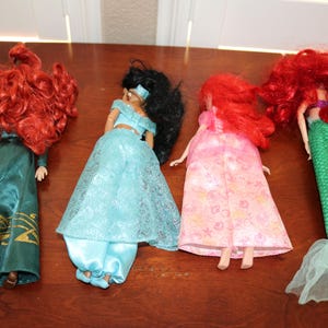 Puede incluir: Cuatro mu&ntilde;ecas se exhiben sobre una superficie de madera. Las mu&ntilde;ecas incluyen una pelirroja con un vestido verde azulado, una mu&ntilde;eca con cabello negro con un atuendo azul, una mu&ntilde;eca con un vestido rosa y una mu&ntilde;eca sirena con cabello rojo y cola verde.