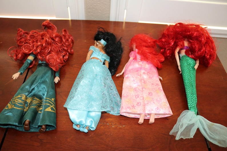 Puede incluir: Cuatro mu&ntilde;ecas se exhiben sobre una superficie de madera. Las mu&ntilde;ecas incluyen una pelirroja con un vestido verde azulado, una mu&ntilde;eca con cabello negro con un atuendo azul, una mu&ntilde;eca con un vestido rosa y una mu&ntilde;eca sirena con cabello rojo y cola verde.