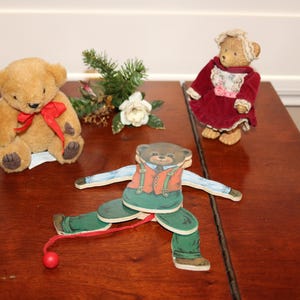 May include: Three teddy bears and a wooden toy bear are arranged on a dark wooden surface. One bear is plush and tan with a red bow. Another is dressed in a red dress. The wooden toy bear has articulated limbs and a red pull string.