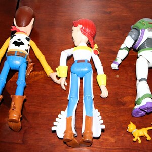 Puede incluir: Tres figuras de personajes de Toy Story: Woody, Jessie y Buzz Lightyear, junto con una peque&ntilde;a figura de gato amarillo. Woody viste un atuendo de vaquero, Jessie tiene un look de vaquera y Buzz lleva un traje espacial.