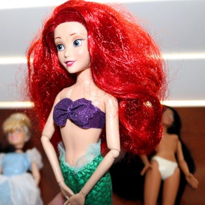 May include: A doll with long, vibrant red hair, blue eyes, and a purple glittery seashell top. The doll features a green, scale-patterned mermaid tail with a light blue tulle trim at the waist. Two other dolls are partially visible in the background.