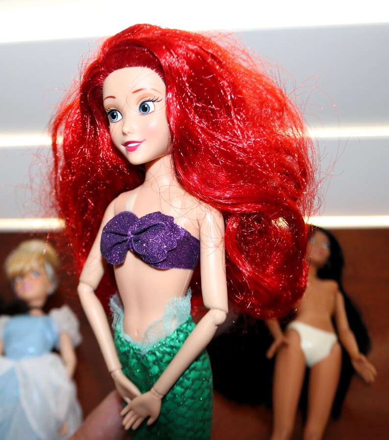 May include: A doll with long, vibrant red hair, blue eyes, and a purple glittery seashell top. The doll features a green, scale-patterned mermaid tail with a light blue tulle trim at the waist. Two other dolls are partially visible in the background.