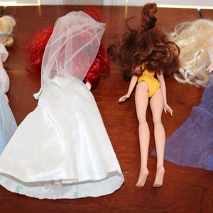 May include: Four fashion dolls are displayed on a wooden surface. The dolls include a blonde doll in a blue gown, a doll in a white dress with a veil, a doll in a yellow swimsuit, and a doll in a blue and white gown.