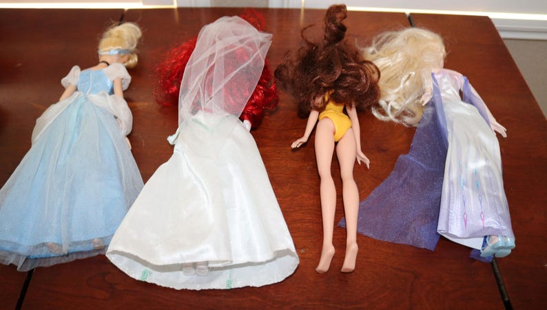 May include: Four fashion dolls are displayed on a wooden surface. The dolls include a blonde doll in a blue gown, a doll in a white dress with a veil, a doll in a yellow swimsuit, and a doll in a blue and white gown.
