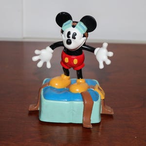 May include: A Mickey Mouse figurine wearing goggles, red shorts, and yellow shoes, standing on a blue and brown base. The figure has black ears, a white face, and white gloves. The base has brown straps.