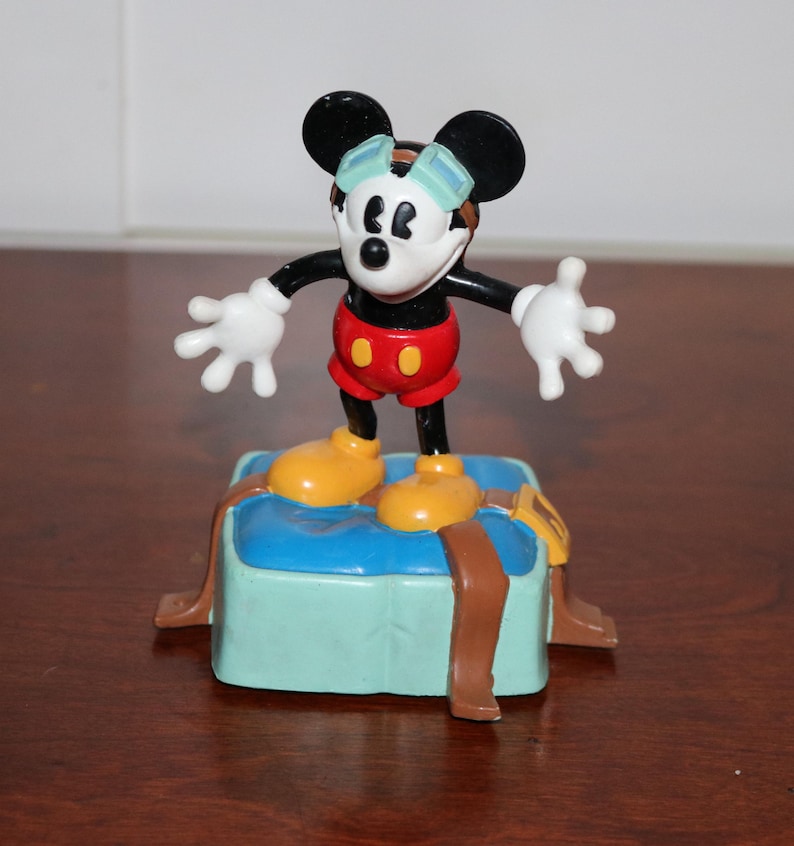 May include: A Mickey Mouse figurine wearing goggles, red shorts, and yellow shoes, standing on a blue and brown base. The figure has black ears, a white face, and white gloves. The base has brown straps.