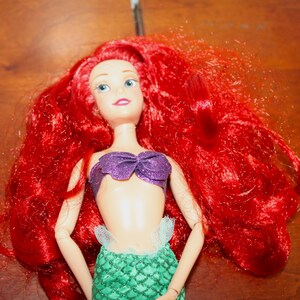 Puede incluir: Una mu&ntilde;eca con cabello rojo, ojos azules y un top morado. La mu&ntilde;eca tiene una cola de sirena verde y es un juguete popular para ni&ntilde;os. El cabello de la mu&ntilde;eca es largo y fluido, y la mu&ntilde;eca sonr&iacute;e.