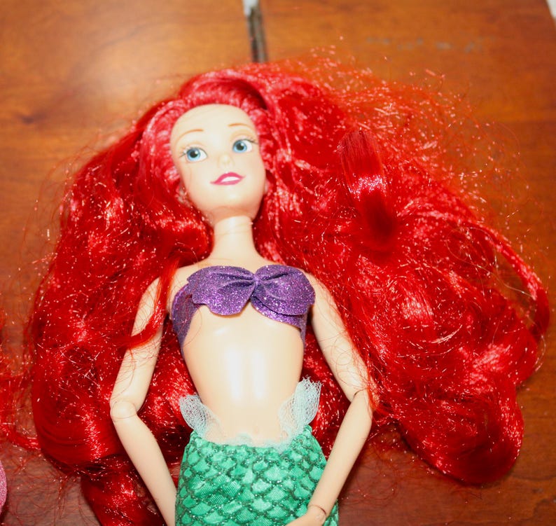 Puede incluir: Una mu&ntilde;eca con cabello rojo, ojos azules y un top morado. La mu&ntilde;eca tiene una cola de sirena verde y es un juguete popular para ni&ntilde;os. El cabello de la mu&ntilde;eca es largo y fluido, y la mu&ntilde;eca sonr&iacute;e.
