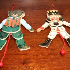 May include: Two wooden jumping jack ornaments featuring a bear and a nutcracker. The bear wears a blue shirt, orange vest, and green trousers. The nutcracker wears a crown, white uniform with red accents, and black boots. Both have red pull strings.