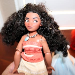 May include: A doll with long, dark, curly hair, brown eyes, and tan skin. The doll wears a coral-colored top with a textured pattern, a light-colored skirt, and a necklace featuring a blue pendant and small white beads around the neck.