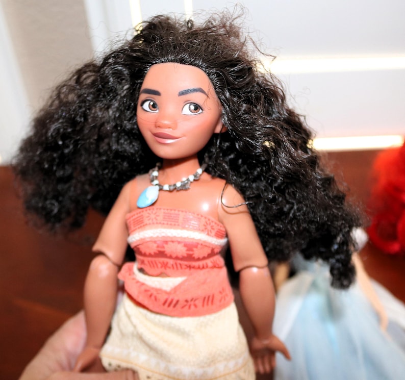 May include: A doll with long, dark, curly hair, brown eyes, and tan skin. The doll wears a coral-colored top with a textured pattern, a light-colored skirt, and a necklace featuring a blue pendant and small white beads around the neck.