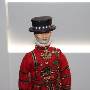 May include: A doll dressed as a Yeoman Warder, wearing a red tunic with black and gold trim, a white ruff collar, and a black top hat with a red, white, and blue band. The figure features a gray beard, blue eyes, and various metallic pins.