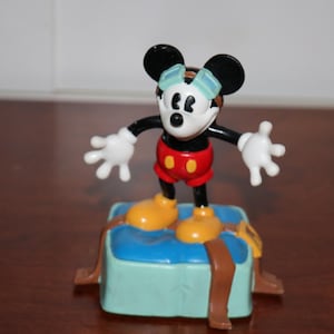 May include: A Mickey Mouse figurine in a classic pose, wearing goggles, red shorts, and yellow shoes. The figure stands on a blue block with brown straps, a collectible Disney item.