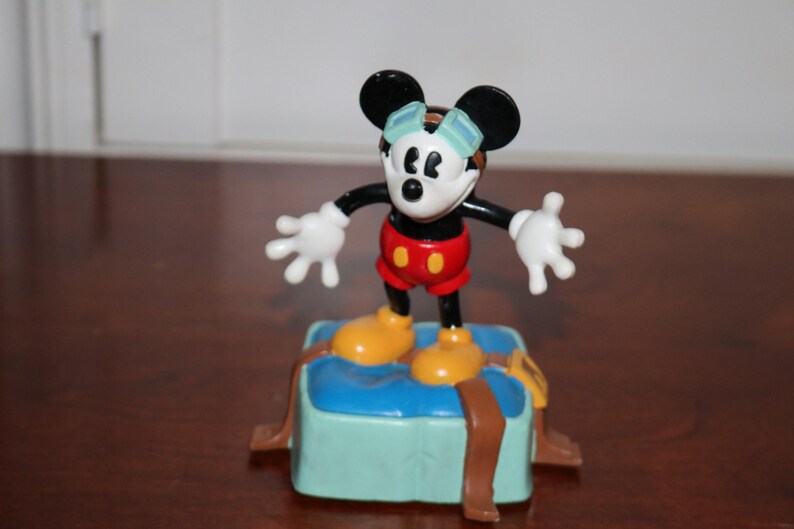 May include: A Mickey Mouse figurine in a classic pose, wearing goggles, red shorts, and yellow shoes. The figure stands on a blue block with brown straps, a collectible Disney item.