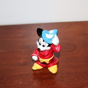 May include: A ceramic figurine of Mickey Mouse dressed as a sorcerer. He is wearing a red robe with a yellow sash and a blue hat with a star on it. He is holding his hat in his right hand and has a mischievous expression on his face.