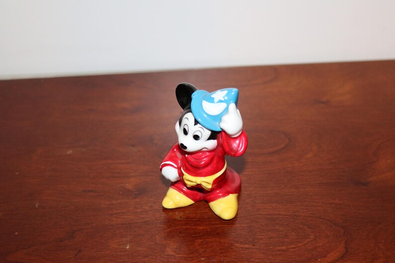 May include: A ceramic figurine of Mickey Mouse dressed as a sorcerer. He is wearing a red robe with a yellow sash and a blue hat with a star on it. He is holding his hat in his right hand and has a mischievous expression on his face.
