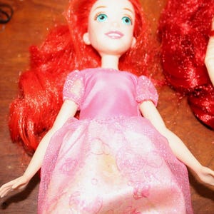 Puede incluir: Una mu&ntilde;eca con cabello rojo brillante y ojos azules, vestida con un vestido rosa con una capa brillante. El vestido tiene un patr&oacute;n detallado. Los brazos de la mu&ntilde;eca est&aacute;n extendidos. La mu&ntilde;eca es un personaje popular de una pel&iacute;cula animada para ni&ntilde;os.