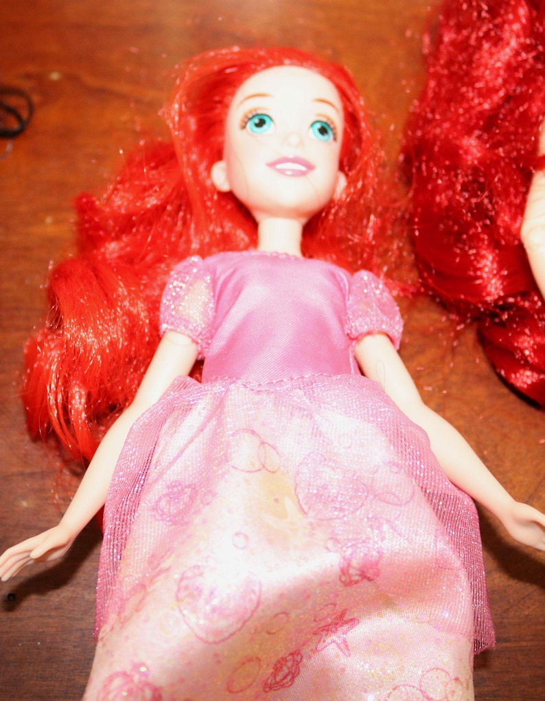 Puede incluir: Una mu&ntilde;eca con cabello rojo brillante y ojos azules, vestida con un vestido rosa con una capa brillante. El vestido tiene un patr&oacute;n detallado. Los brazos de la mu&ntilde;eca est&aacute;n extendidos. La mu&ntilde;eca es un personaje popular de una pel&iacute;cula animada para ni&ntilde;os.