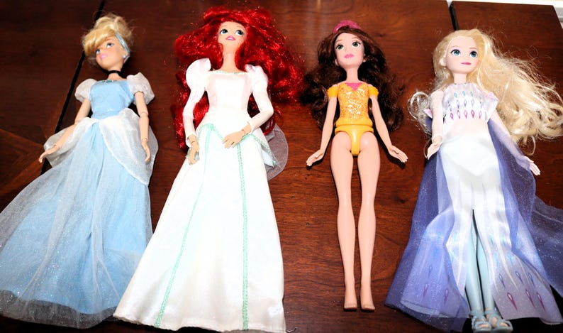 May include: Four dolls of Disney princesses are displayed. Cinderella wears a blue gown, Ariel has a white dress and red hair, Belle is in a yellow outfit, and Elsa has a blue and white dress.