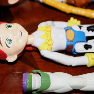 Puede incluir: Primer plano de varias figuras de acci&oacute;n de personajes de Toy Story. Jessie, con cabello rojo y un atuendo blanco con detalles amarillos y azules, est&aacute; en primer plano. Otros personajes, incluidos Woody y Buzz Lightyear, son parcialmente visibles.