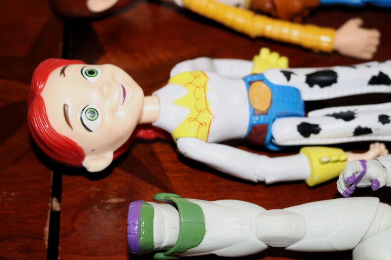 Puede incluir: Primer plano de varias figuras de acci&oacute;n de personajes de Toy Story. Jessie, con cabello rojo y un atuendo blanco con detalles amarillos y azules, est&aacute; en primer plano. Otros personajes, incluidos Woody y Buzz Lightyear, son parcialmente visibles.