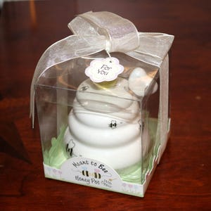May include: A white ceramic honey pot with a bee design, presented in a clear box with a ribbon. The packaging includes the text "Meant to Bee Honey Pot With Dipper" and a "For You" tag.