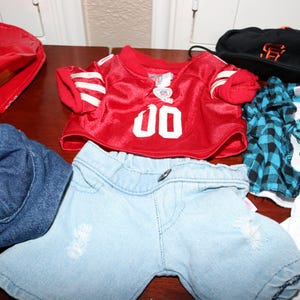 May include: A collection of children's clothing items, including a red jersey with the number 00, denim shorts, a black baseball cap, and a blue and black plaid shirt. Other items include a red patent leather bag and a white t-shirt with a dinosaur graphic.