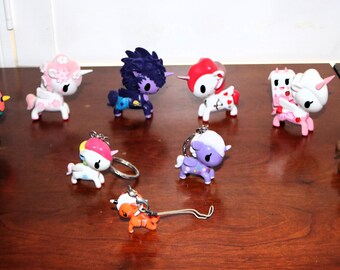 May include: A collection of ten small unicorn figurines and keychains in various colors including green, pink, purple, red, and brown. The figures feature distinct designs, some with wings, horns, and patterned markings, arranged on a dark wooden surface.