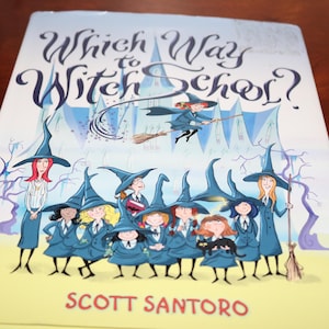 May include: A children's book titled "Which Way to Witch School?" features illustrations of witches in blue uniforms and hats. The cover art includes the author's name, Scott Santoro, and a whimsical depiction of witches flying on broomsticks.